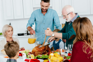 grandfather slicing baked turkey at served table while family celebrating thanksgiving