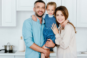 Fototapeta premium portrait of happy family holding and embracing little daughter and looking at camera at kitchen