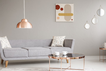 Real photo of a simple sofa, copper lamp and table, and painting on a wall in a living room interior