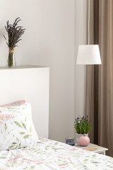 Lamp above cabinet with plant next to bed with sheets in bedroom interior with lavender flowers. Real photo