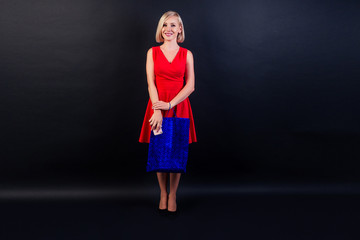 attractive blond woman in red dress holds a leather blue bag in high heels full-length studio shot in black background. black Friday shopping sale concept