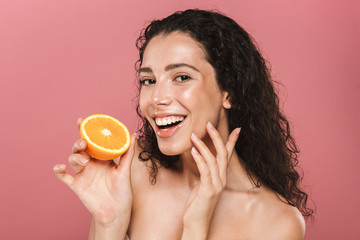 Portrait of brunette half naked woman with long hair smiling and holding piece of orange, isolated over pink background