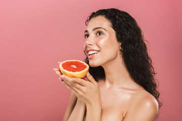 Beauty photo of caucasian woman with long hair smiling and holding piece of grapefruit, isolated over pink background