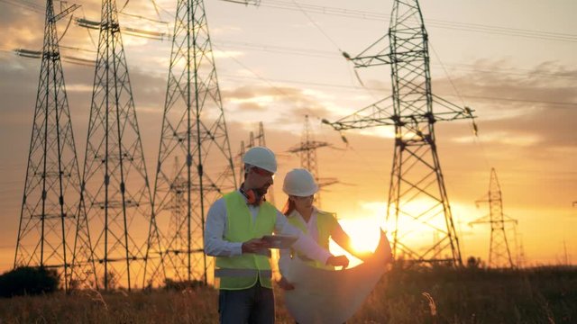 Two engineers in uniform work near power lines. Sunset background.