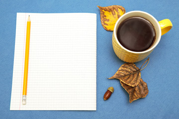 A sheet of paper with a pencil and a cup of tea on a blue background, next to the autumn yellow leaves, top view. Autumn composition. Space for text.