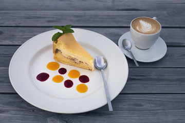 piece of appetizing cheesecake with drops of jam on a plate and a cup of coffee on a wooden background