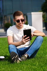 Obraz premium Young handsome guy with sunglasses using tablet and headphones, drinking coffee to go, sitting on grass and enjoying nice sunny day by modern building. Male model photo-shoot, urban fashion lifestyle.