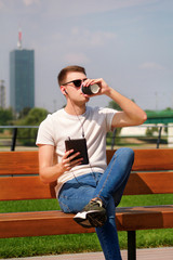 Fototapeta premium Handsome boy wearing sunglasses using tablet and headphones, sitting on a bench and drinking coffee to go. Handsome student immersed in social media. Background scenery bridge, river and green grass.