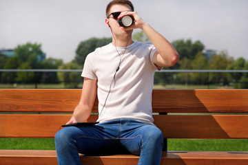 Obraz premium Handsome boy wearing sunglasses using tablet and headphones, sitting on a bench and drinking coffee to go. Handsome student immersed in social media. Background scenery bridge, river and green grass.