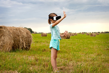 Naklejka na ściany i meble A little girl in virtual reality glasses, standing in the field and playing.