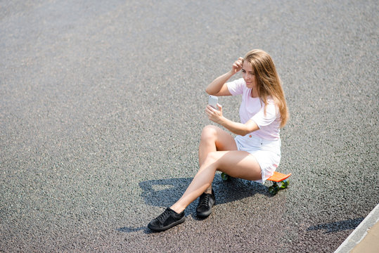 Young Beautiful Smiling Girl Making Selfie with Smartphone while Sitting on the Skateboard on the Road