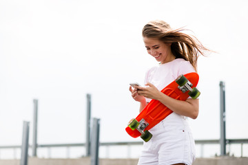 Young Beautiful Smiling Blonde Girl Using Smartphone while Sitting on the Skateboard