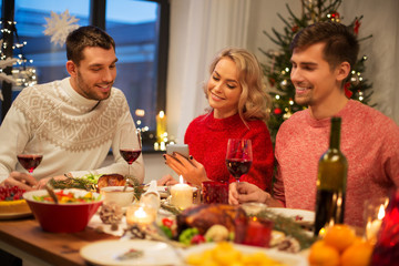 holidays and celebration concept - happy friends with smartphone having christmas dinner at home