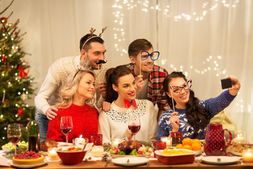 holidays and celebration concept - happy friends with party props taking selfie by smartphone at home christmas dinner