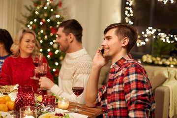 holidays, communication and celebration concept - happy young man calling on smartphone and having christmas dinner with friends at home
