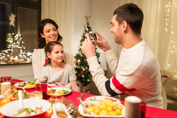 holidays, family and celebration concept - happy mother, father and little daughter having christmas dinner and taking picture by smartphone at home