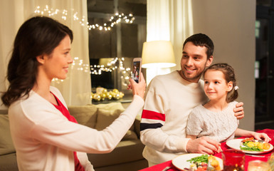 holidays, family and celebration concept - happy mother, father and little daughter having christmas dinner and taking picture by smartphone at home