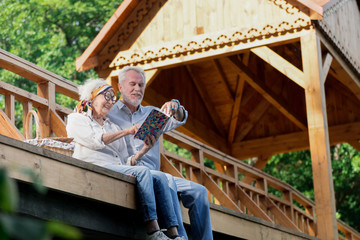 Memorable notebook. Positive elderly couple sitting on the wooden bridge and looking at the notebook