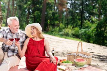 Having a picnic. Cheerful elderly couple having little picnic in the forest and drinking red wine