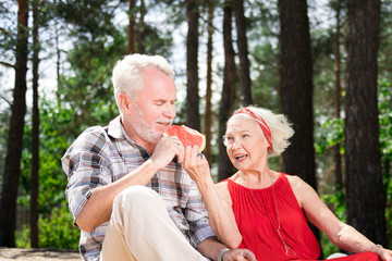 Eating watermelon. Kind reliable senior woman feeding her husband with tasty watermelon while having a picnic with him