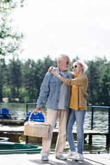 Hugging on pier. Cheerful senior man hugging his wife and holding a basket while standing on the pier