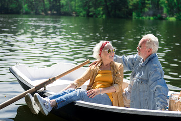 Feeling relaxed. Calm curious senior woman smiling to her serious husband while sitting in the boat with him