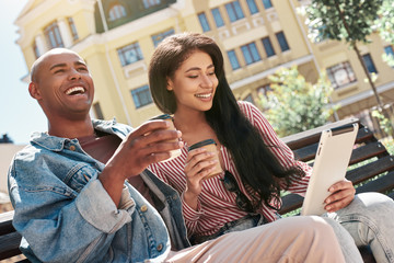 Using gadgets. Young diverse couple sitting on bench on the city street holding cups drinking hot coffee woman browsing internet on digital tablet laughing happy