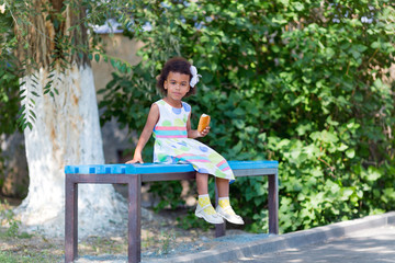 Naklejka premium A black girl is sitting on a bench with bread. A little girl is eating a patty in the city park.