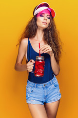 Sensual young woman in summer outfit, cap holding jar with fresh beverage and while standing on bright yellow background