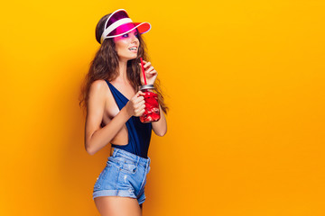 Sensual young woman in summer outfit, cap holding jar with fresh beverage and while standing on bright yellow background