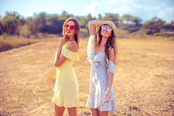 two pretty womem walking in the field dressed in summer dresses, hat, sunglasses and looking at camera
