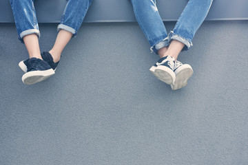 partial view of couple of legs in jeans and sneakers sitting on grey wall