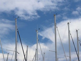 Masts of sailing yachts against the blue sky with white clouds. Yacht in the marina during the morning dawn sailing past the moored sailing yachts. Marine life style. Romantic and extreme rest on the 