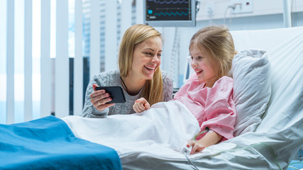 Cute Little Girl Lies on a Bed in the Children's Hospital, Her Mother Sits Beside, They Watch Cartoons/ Funny Videos on Smartphone. Modern Pediatric Ward.