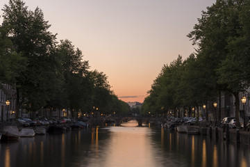 Fototapeta premium Zachód słońca, patrząc na kanał w Amsterdamie. Niebo jest pomarańczowe, a światła uliczne odbijają się od spokojnej wody