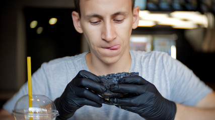 A young man sits in a restaurant and looks carefully at the burger who holds in his hands