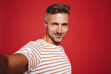 Beautiful guy 30s in striped t-shirt smiling while taking selfie photo, isolated over red background