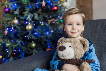 adorable happy child hugging teddy bear and smiling at camera while sitting on sofa at christmas time