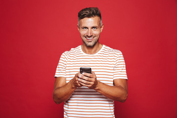 Image of joyful man in striped t-shirt smiling and holding cell phone, isolated over red background