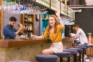 Oh hello. Positive young girl sitting at the bar counter and feeling excited while waving to friends