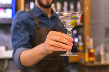 Tasty alcohol. Careful professional barman holding a glass of delicious alcohol with an olive