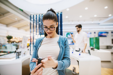 Beautiful modern girl is testing new telephone in tech store while her boyfriend is looking around in a store.