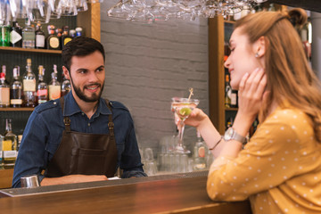Drinking and talking. Relaxed young girl sitting at the bar counter with glass of alcohol and talking to smiling barman