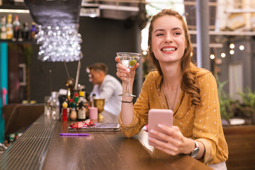 Girl with smartphone. Cheerful young girl sitting at the bar counter with glass of alcohol and holding her smartphone