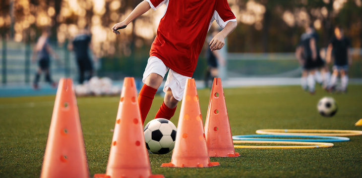 Fototapeta Soccer Drills: The Slalom Drill. Youth soccer practice drills. Young football player training on pitch. Soccer slalom cone drill. Boy in red soccer jersey shirt running with ball between cones