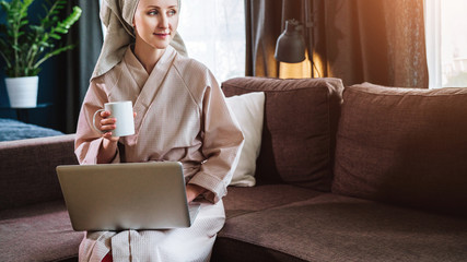 Morning. Young woman in bathrobe and towel on her head sits in room on couch, drinks coffee and...