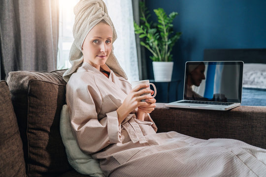 Young woman in bathrobe and towel on head sitting in room on couch,drinking coffee,working on laptop.Morning, girl after shower drinks tea, uses computer. Freelancer works home, cozy home environment.