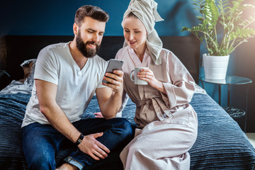 Morning.Young couple,woman after shower in bathrobe and towel on her head, sitting in bedroom on bed.Man shows information to woman on smartphone, girl looks at screen, drinks coffee. Social networks.