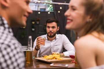 Odd friend. Calm young man thoughtfully drinking his alcohol and looking at two friends flirting
