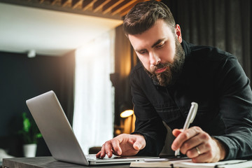Businessman is standing near computer, working on laptop, making notes in notebook. Man watching webinar, learning.